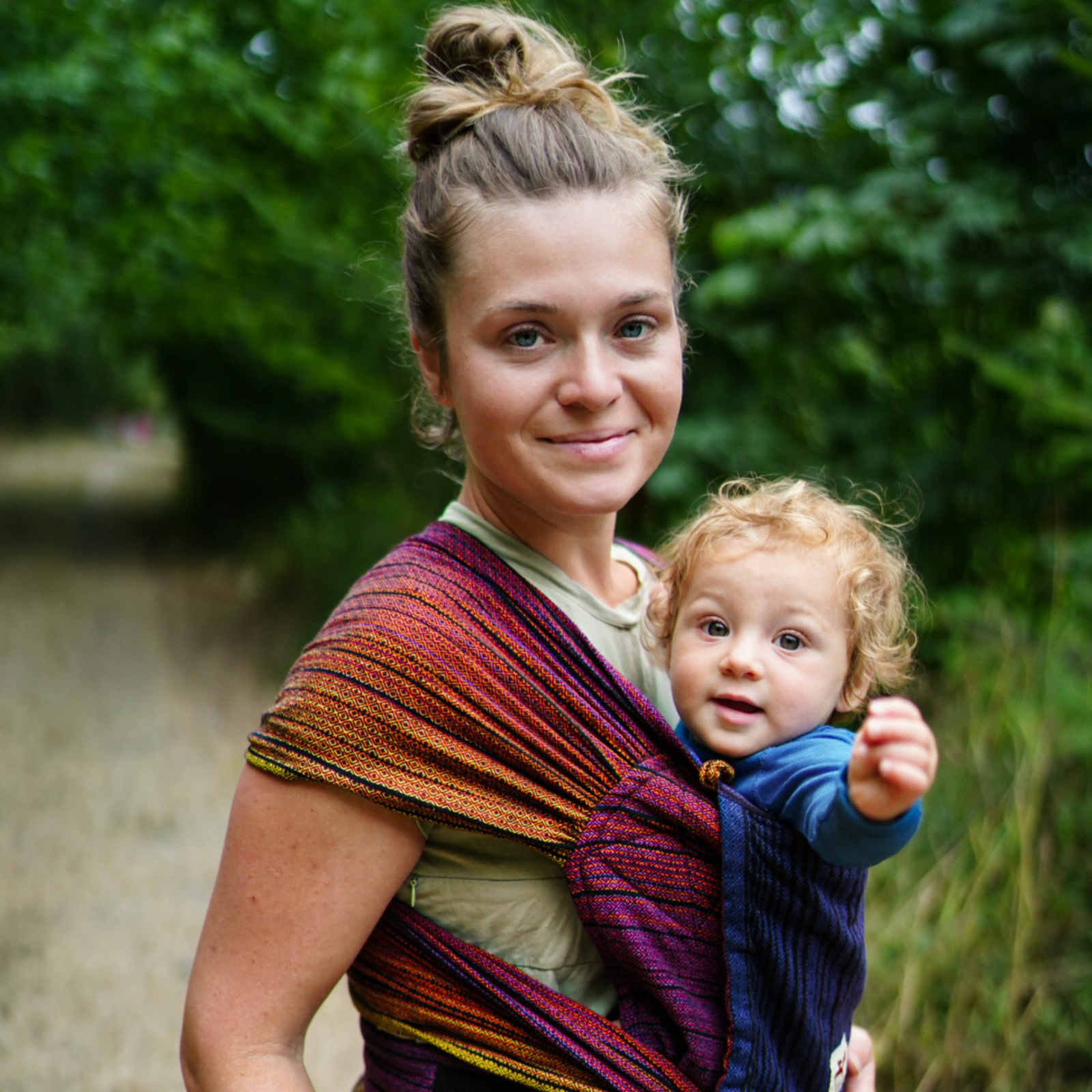 Mutter lächelt, Baby in der Trage WrapMysol Twilight schaut neugierig und streckt die Hand zum Fotografen